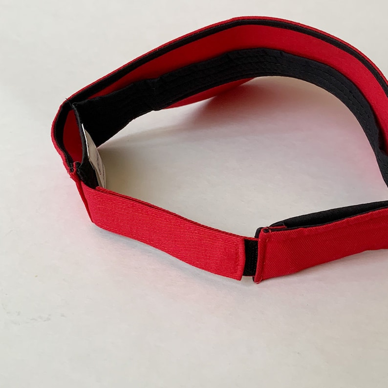 Op de afbeelding: Een rode visor met een zwarte verstelbare band. De visor is gemaakt van een zacht, flexibel materiaal.