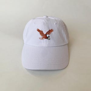 May include: White baseball cap with an embroidered brown eagle design.