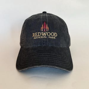 Puede incluir: Una gorra de béisbol de algodón negro, desgastada, con un logotipo bordado de tres secuoyas y el texto "Redwood National Park" en dorado.