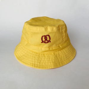 May include: A yellow bucket hat with a brown pretzel embroidered on the front.
