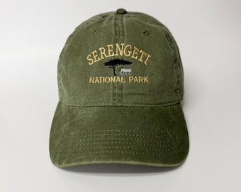May include: An olive green baseball cap featuring the embroidered text "SERENGETI NATIONAL PARK" in gold. The design includes a tree and a zebra. The cap has a curved peak and an adjustable strap at the back.