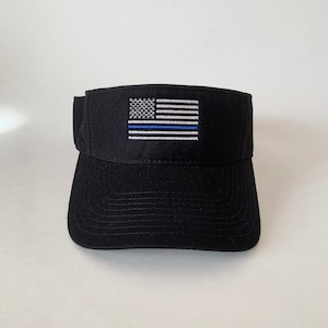 May include: Black visor featuring an embroidered American flag with a blue line. The visor has a curved brim and a structured design. The flag design includes white stripes, a blue field with white stars, and a blue stripe.