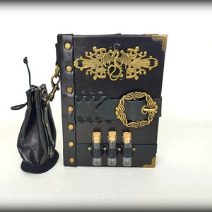 Puede incluir: Libro de fantasía de cuero negro con detalles dorados, un emblema de dragón y un cierre de hebilla. Incluye una pequeña bolsa con cordón y tres viales de vidrio con tapones de corcho. El libro tiene esquinas y remaches de latón.