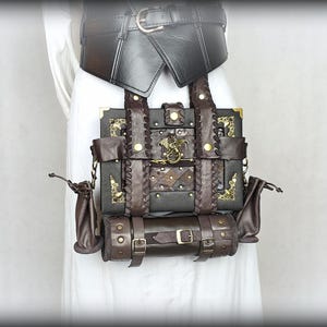 Könnte beinhalten: Eine rechteckige, dunkelbraune Tasche in Buchform mit geflochtenen Lederriemen, goldfarbenen Metallecken und einem zentralen Drachenemblem. Darunter befindet sich eine zylindrische Lederrolle mit Schnallen, flankiert von zwei kleinen Beuteln.