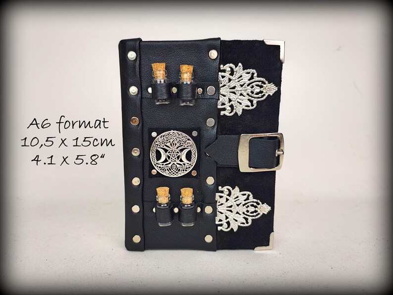 May include: Black leather A6 format journal with silver accents. The journal features a decorative silver emblem, two small glass bottles with cork stoppers, and a black buckle closure. The dimensions are 4.1 x 5.8 inches.