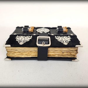 May include: A black leather-bound journal with silver corner protectors and a buckle closure. The journal features ornate silver embellishments and a black suede panel. The aged pages are visible, and the journal is secured with a black leather strap.