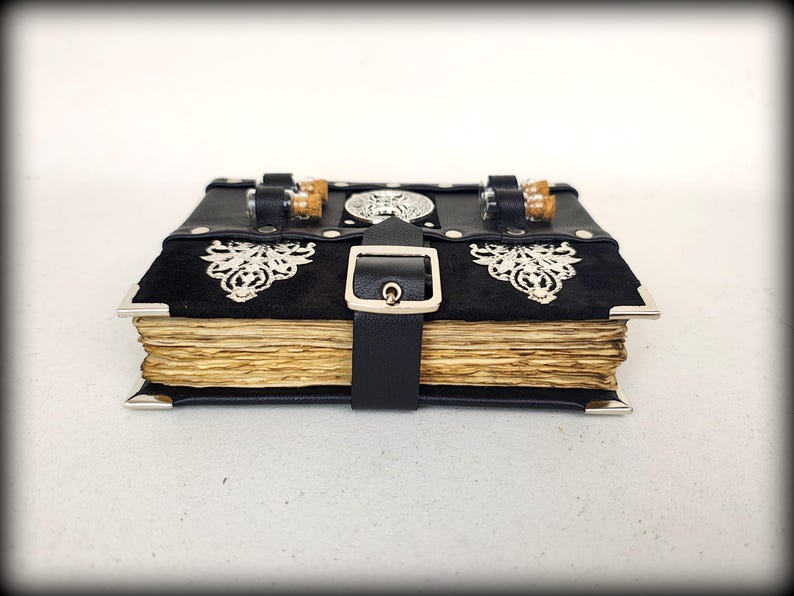 May include: A black leather-bound journal with silver corner protectors and a buckle closure. The journal features ornate silver embellishments and a black suede panel. The aged pages are visible, and the journal is secured with a black leather strap.