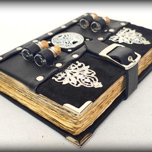 May include: A handcrafted black leather journal with aged paper pages. The journal features silver corner protectors, decorative metal accents, and a black strap with a silver buckle. Small glass vials are attached to the top.