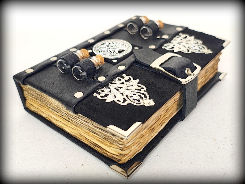 May include: A handcrafted black leather journal with aged paper pages. The journal features silver corner protectors, decorative metal accents, and a black strap with a silver buckle. Small glass vials are attached to the top.