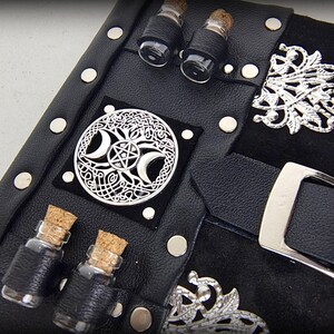 May include: Black leather journal with silver accents, including a buckle, decorative filigree, and a circular emblem featuring a crescent moon and pentagram design. Small glass vials with cork stoppers are attached.