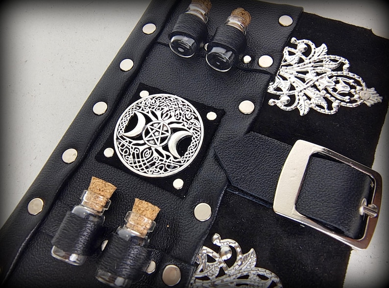 May include: Black leather journal with silver accents, including a buckle, decorative filigree, and a circular emblem featuring a crescent moon and pentagram design. Small glass vials with cork stoppers are attached.