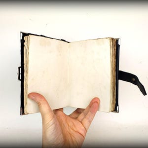 May include: An open, antique-style journal with aged, cream-colored pages. The journal has a dark blue cover with metal corner protectors and a black leather strap closure. The pages are held open by a hand.
