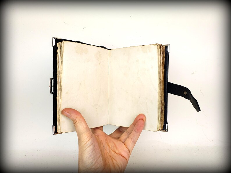 May include: An open, antique-style journal with aged, cream-colored pages. The journal has a dark blue cover with metal corner protectors and a black leather strap closure. The pages are held open by a hand.