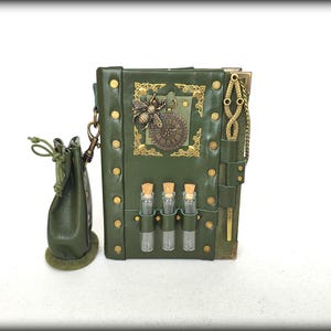 Può includere: Un diario rilegato in pelle verde scuro con un emblema in ottone raffigurante un'ape e un quadrante d'orologio, borchie in metallo dorato e tre piccole fiale di vetro con tappi di sughero sul davanti. Una pochette in pelle verde è agganciata al lato.