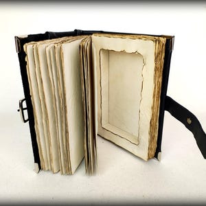 May include: An open, antique-style book with aged, cream-colored pages. The book has a black leather cover and a metal clasp. The pages have a distressed, burnt-edge appearance, and the book is propped open with a black leather stand.