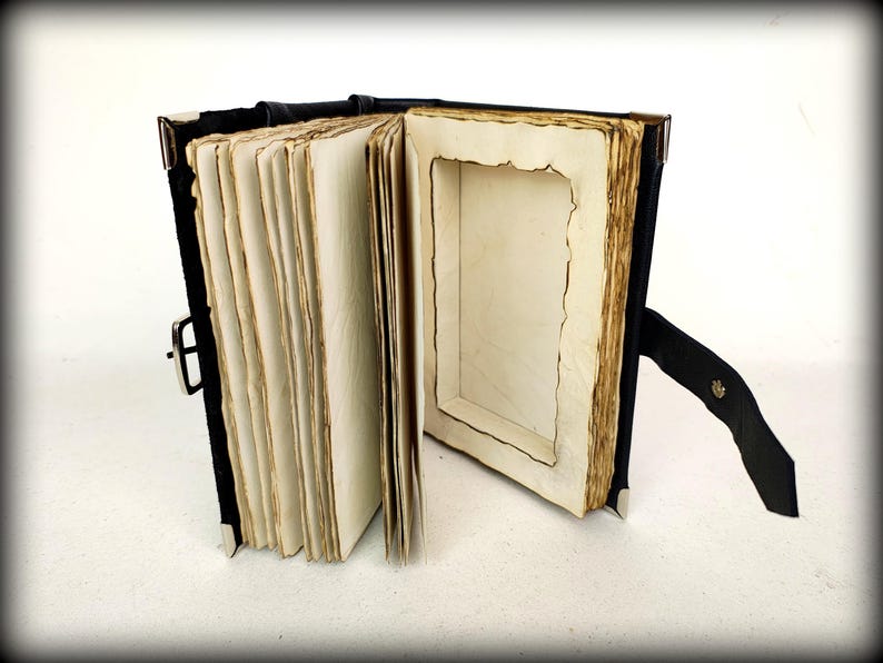 May include: An open, antique-style book with aged, cream-colored pages. The book has a black leather cover and a metal clasp. The pages have a distressed, burnt-edge appearance, and the book is propped open with a black leather stand.