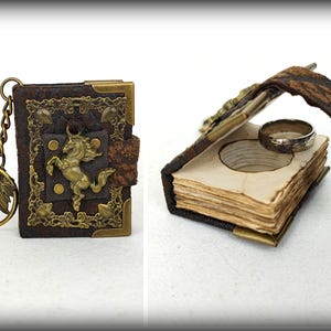 Puede incluir: Dos libros de cuero en miniatura. Uno es un llavero con un emblema de caballo dorado y un dije de hoja. El otro está abierto, revelando un anillo en el interior. Ambos libros tienen adornos de metal en las esquinas y cierres.