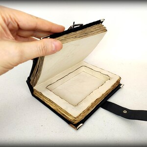 May include: A handcrafted, open book with aged, cream-colored pages and a black leather cover. The book has a recessed interior and a black leather strap with a metal clasp. The pages have a rough, uneven edge.