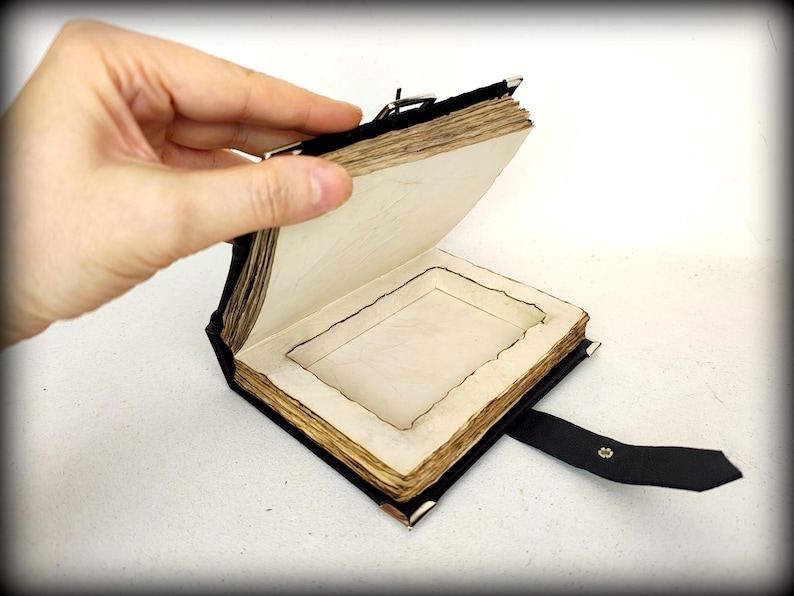 May include: A handcrafted, open book with aged, cream-colored pages and a black leather cover. The book has a recessed interior and a black leather strap with a metal clasp. The pages have a rough, uneven edge.
