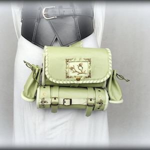 May include: A light green multi-part belt bag featuring a rectangular main pouch with a central metallic branch emblem, a cylindrical lower compartment with two buckle straps, and two small side pouches with drawstring closures, worn over a black belt.