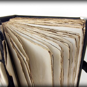May include: A partially open, antique-style book with aged, cream-colored pages. The book has a black leather cover and silver corner protectors. The pages have a distressed, weathered appearance, suggesting age and use. The book is open, revealing the texture of the paper.