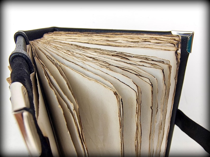 May include: A partially open, antique-style book with aged, cream-colored pages. The book has a black leather cover and silver corner protectors. The pages have a distressed, weathered appearance, suggesting age and use. The book is open, revealing the texture of the paper.
