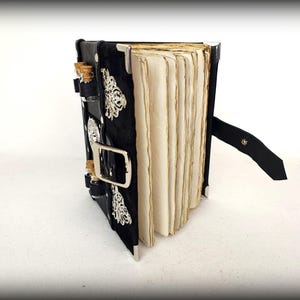 May include: A black, handmade journal with aged paper pages. The cover is adorned with silver embellishments and small glass vials with cork stoppers. A black leather strap with a buckle secures the journal.