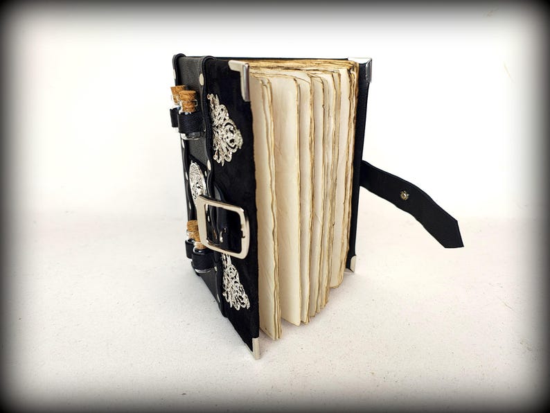 May include: A black, handmade journal with aged paper pages. The cover is adorned with silver embellishments and small glass vials with cork stoppers. A black leather strap with a buckle secures the journal.