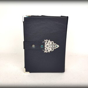 May include: A dark blue leather-bound journal with silver corner protectors and a decorative silver clasp. The clasp features an ornate design with a small green gem in the center. A dark blue strap secures the journal.