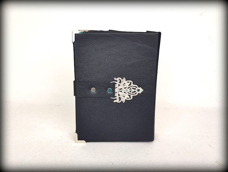May include: A dark blue leather-bound journal with silver corner protectors and a decorative silver clasp. The clasp features an ornate design with a small green gem in the center. A dark blue strap secures the journal.