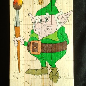 Può includere: Un puzzle in legno con un folletto verde che tiene un pennello con una punta a forma di fiamma. Il folletto ha una barba bianca, un cappello verde con un campanello e indossa una cintura marrone. Il puzzle è composto da 16 pezzi.