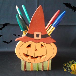 Puede incluir: Un porta lápices de madera con forma de calabaza con un sombrero de bruja en la parte superior. El sombrero es rojo con una banda negra y una hebilla. La calabaza es naranja con una cara de calabaza tallada. Hay cinco bolígrafos y lápices en el porta lápices.