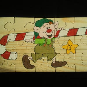 Puede incluir: Un rompecabezas de madera con un duende de dibujos animados sosteniendo un bastón de caramelo. El duende lleva un sombrero verde y una camisa a rayas rojas y blancas. El rompecabezas está sobre un fondo marrón.