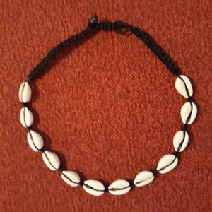 May include: A black cord necklace with white seashells strung on it. The necklace is on a red fabric background.