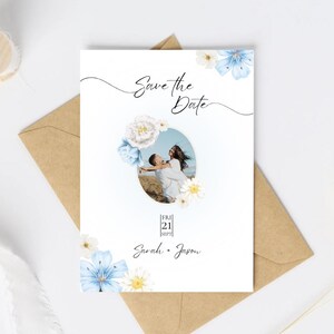 May include: A white wedding invitation with the text "Save the Date" and a photo of a couple. The invitation is decorated with blue and white floral accents and rests on a brown envelope.