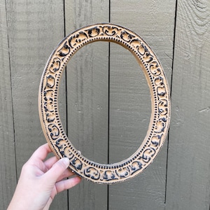 May include: Oval-shaped, cream-colored picture frame with intricate black detailing. The frame is held by a hand against a weathered, gray-green wooden background. The frame's design features repeating floral patterns.