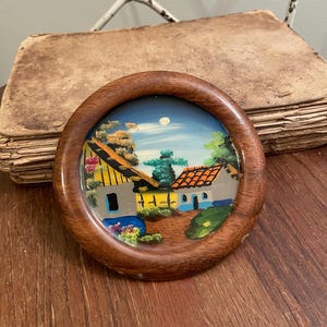 May include: A round, wooden framed painting depicting a village scene with houses, trees, and a blue sky with a moon. The painting has a brown wooden frame. The artwork is on a wooden surface.