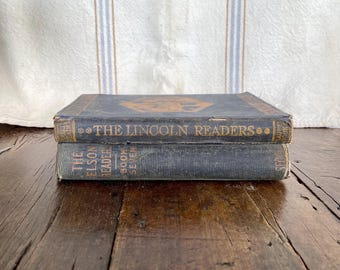 May include: Two antique books stacked on a wooden surface. The top book is titled "The Lincoln Readers" in gold lettering, with a decorative emblem. The bottom book is titled "The Elson Readers Book Seven". The books have a distressed, aged appearance.