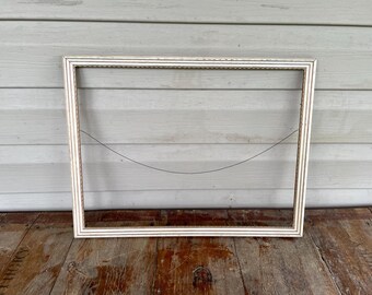 May include: A rectangular, empty picture frame with a white finish and a decorative edge. The frame is set against a weathered, light-colored wooden background and a dark wooden surface.