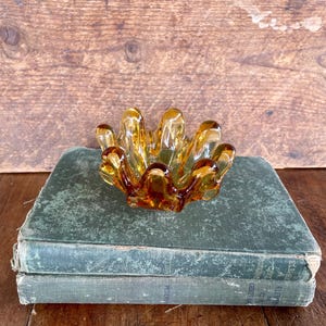May include: A vintage amber glass ashtray with a unique, organic shape, resting on a stack of antique books with faded green covers. The ashtray's design features multiple rounded sections, creating a flower-like appearance. The background is a rustic wooden surface.