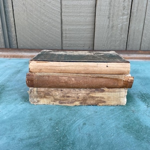 May include: A stack of three antique books with worn, aged covers. The top book has a dark green cover, the middle book has a brown spine, and the bottom book has a light brown, distressed cover. The books are stacked on a teal surface.