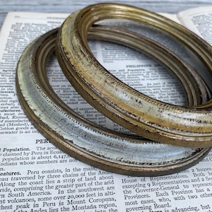 May include: Two oval picture frames with a distressed finish. The top frame is gold with dark brown accents, while the bottom frame features a weathered white and gold appearance. Both frames have a beaded inner edge detail.