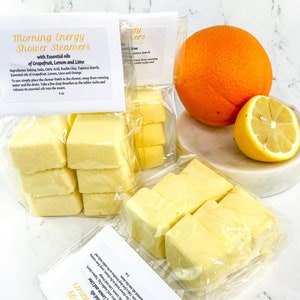 May include: Two clear plastic bags filled with yellow shower steamers, labeled "Morning Energy Shower Steamers with Essential Oils of Grapefruit, Lemon and Lime".  The bags are next to a whole orange and a lemon on a white marble surface.