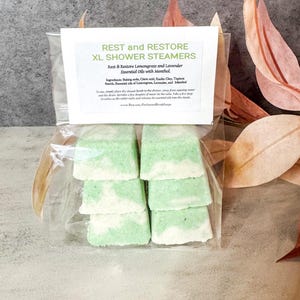 May include: A clear plastic bag containing six green and white shower steamers. The label reads "REST and RESTORE XL SHOWER STEAMERS" with ingredients listed. The background is a neutral gray with fall-colored leaves.