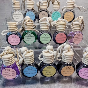 May include: A collection of small, round glass car air freshener bottles with wooden caps and hanging ropes. Each bottle contains colored liquid and a label with scent names like Vanilla Veil, Pine Forest, Garden Therapy, and Pumpkin Souffle.