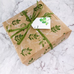 May include: A brown gift box wrapped in paper with a green leaf pattern. The box is tied with a green ribbon and has a white gift tag with the words "To" and "From" written on it.
