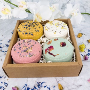 May include: A set of four bath bombs in a brown cardboard box. The bath bombs are round and have different colors and floral designs. The bath bombs are arranged in a square pattern in the box.