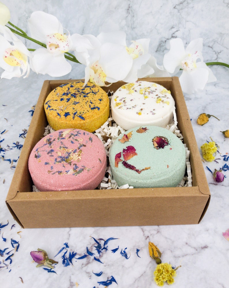 May include: A set of four bath bombs in a brown cardboard box. The bath bombs are round and have different colors and floral designs. The bath bombs are arranged in a square pattern in the box.
