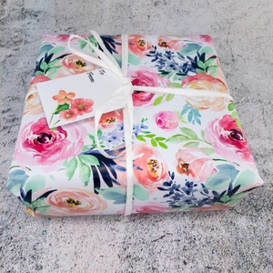 May include: A gift box wrapped in white paper with a floral pattern in shades of pink, peach, blue, and green. The box is tied with a white ribbon and has a white gift tag that says "To" and "From:".