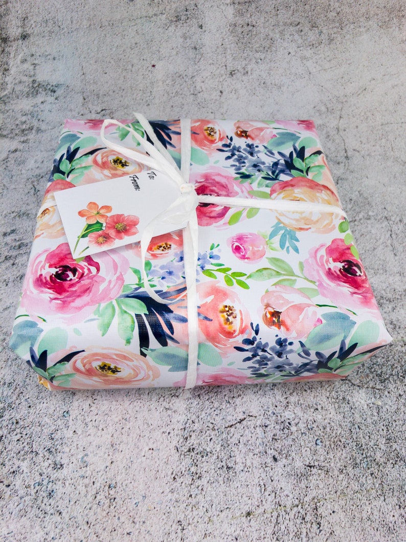 May include: A gift box wrapped in white paper with a floral pattern in shades of pink, peach, blue, and green. The box is tied with a white ribbon and has a white gift tag that says "To" and "From:".
